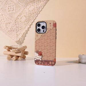 May include: A tan phone case featuring a Hello Kitty design with autumn leaves. The case has a patterned background and a decorative border. The case is designed to fit a smartphone.