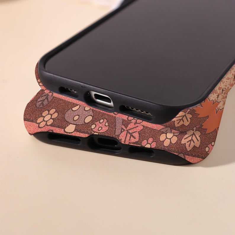 May include: Two black smartphones stacked on top of each other. The bottom phone case has a brown background with a pattern of mushrooms, leaves, and flowers in shades of orange, pink, and white.