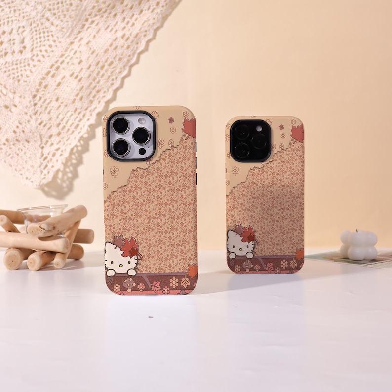 May include: Two phone cases with a Hello Kitty design. The cases are tan with a floral pattern and a Hello Kitty graphic at the bottom. The cases have a brown border with fall leaves. The phone cases are different sizes.