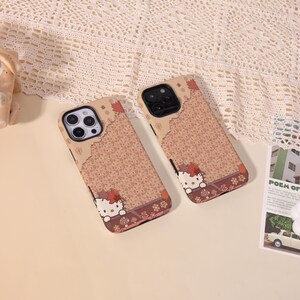 May include: Two phone cases with a Hello Kitty design. The cases have a brown floral pattern with a Hello Kitty graphic at the bottom. The top of the case is a lighter brown color. The cases are on a beige surface.