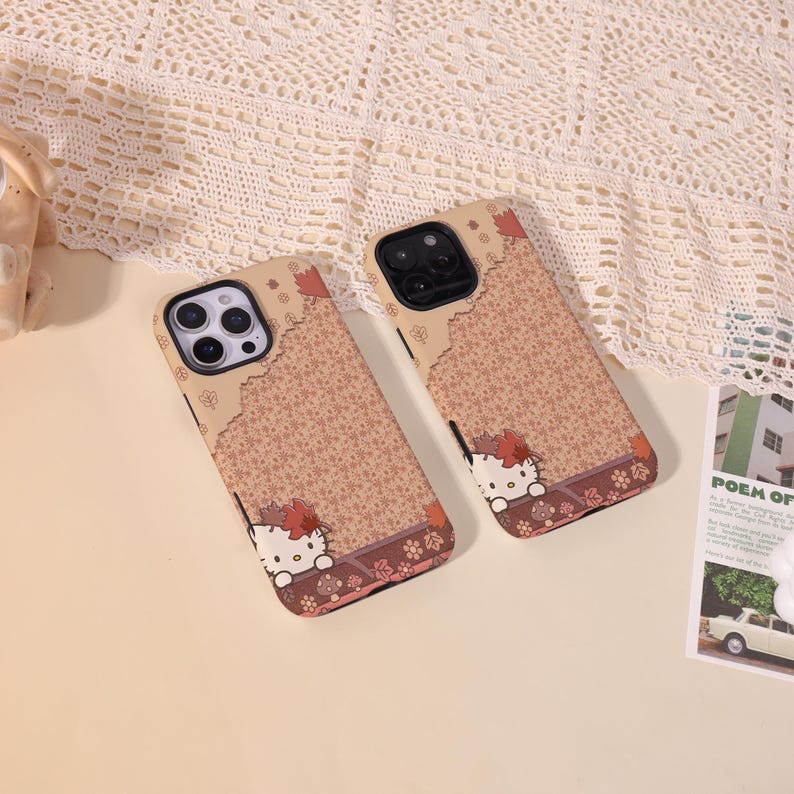 May include: Two phone cases with a Hello Kitty design. The cases have a brown floral pattern with a Hello Kitty graphic at the bottom. The top of the case is a lighter brown color. The cases are on a beige surface.