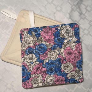 May include: Two square quilted pot holders. One features a vibrant floral pattern of blue, pink, and white roses. The other is a solid, plain cream-colored fabric with a white hanging loop attached to the corner for convenient storage in the kitchen.