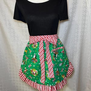 May include: A green Christmas-themed apron with a red and white striped waistband and trim. The apron features a festive pattern with holiday characters and candy canes. The apron is tied in a bow at the waist.