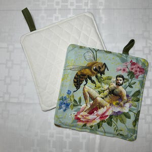 May include: Two square pot holders. One is white with a quilted diamond pattern and an olive green loop. The other has a light blue background with a bee and a man in a flower design, with an olive green loop.
