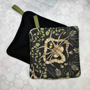 May include: Two square pot holders featuring a black background with a repeating pattern of beige, multi-legged creatures, green leaves, and small yellow flowers. Each pot holder has a solid black reverse side and a small olive green fabric hanging loop.