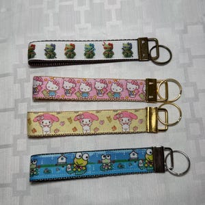 May include: Four keychains with patterned ribbons. The top keychain has a white ribbon with green frog designs. The second keychain has a pink ribbon with Hello Kitty designs. The third keychain has a yellow ribbon with My Melody designs. The bottom keychain has a blue ribbon with frog designs.