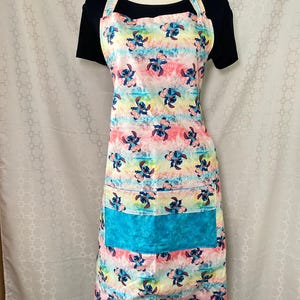 May include: A colorful apron featuring a repeating pattern of a blue cartoon character on a pastel background of pink, yellow, and blue. The apron has a turquoise pocket and neck and waist ties.