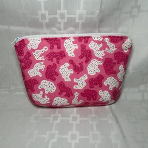 May include: A pink zippered pouch with a repeating pattern of white and magenta animal shapes. The pouch has a white zipper and a rounded rectangular shape. The background is a white fabric with a geometric pattern.