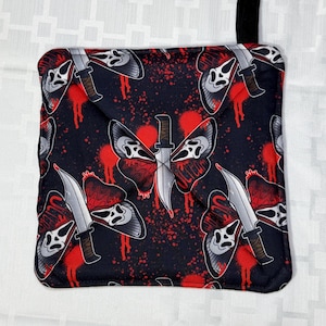 May include: Black pot holder with a repeating pattern of a white ghost face with a knife through a butterfly. The design includes red blood splatter and the word "SCREAM". A black loop is attached to the top corner.
