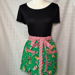 May include: A green apron with a Christmas-themed pattern featuring cartoon characters, candy canes, and snowflakes. The apron has a red and white striped waistband and ties, and two pockets. The apron is displayed on a mannequin.