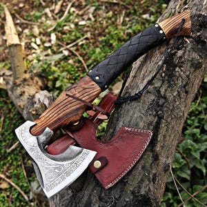 Peut inclure: Une hache artisanale avec un manche en bois et une lame argentée. Le manche est enveloppé de cuir noir. La hache repose sur une branche d'arbre avec un étui en cuir marron.