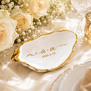 May include: A white oyster shell trinket dish with a gold-painted rim. The interior features gold lettering that reads a + m and the date 2026.6.18, centered in a decorative script font with flourishes on either side of the letters.