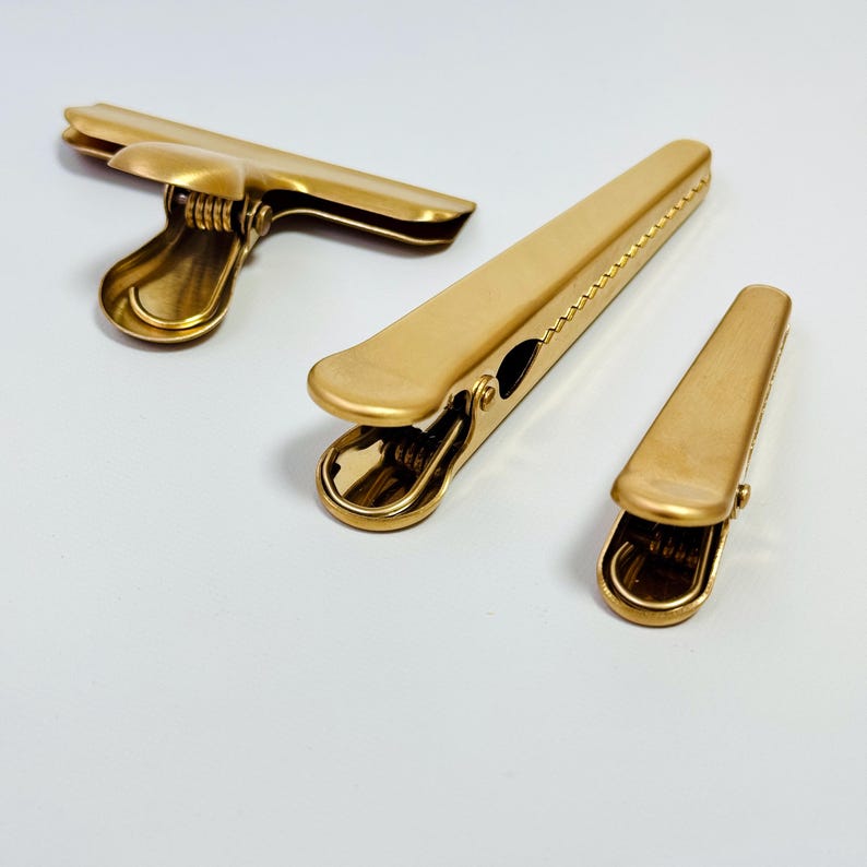 K&ouml;nnte beinhalten: Drei goldfarbene Metall-Ordnerklammern in verschiedenen Gr&ouml;&szlig;en sind auf einer wei&szlig;en Oberfl&auml;che angeordnet. Die Klammern haben eine glatte, matte Oberfl&auml;che und ein klassisches Design mit Federmechanismus f&uuml;r sicheren Verschluss. Die gr&ouml;&szlig;te Klammer ist ge&ouml;ffnet, die anderen beiden sind geschlossen.