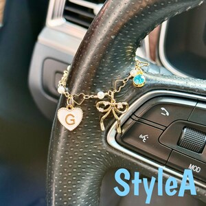 May include: Gold-toned car charm with a white heart pendant featuring the letter "G", a bow, and a blue gem. The charm is attached to a car steering wheel. The text "StyleA" is visible in the image.