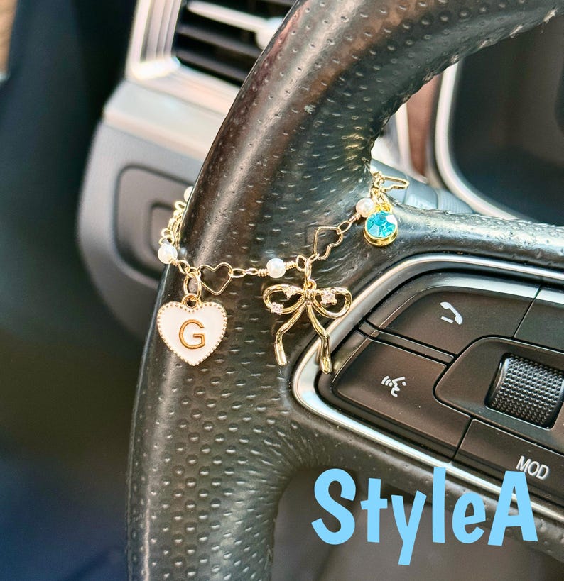 May include: Gold-toned car charm with a white heart pendant featuring the letter "G", a bow, and a blue gem. The charm is attached to a car steering wheel. The text "StyleA" is visible in the image.