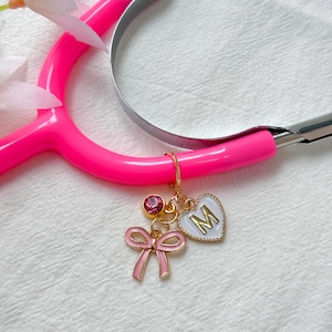 Puede incluir: Un estetoscopio rosa brillante con un dije dorado. El dije incluye un lazo rosa, una gema rosa y un coraz&oacute;n blanco con la letra "M". El estetoscopio est&aacute; sobre una superficie blanca texturizada.