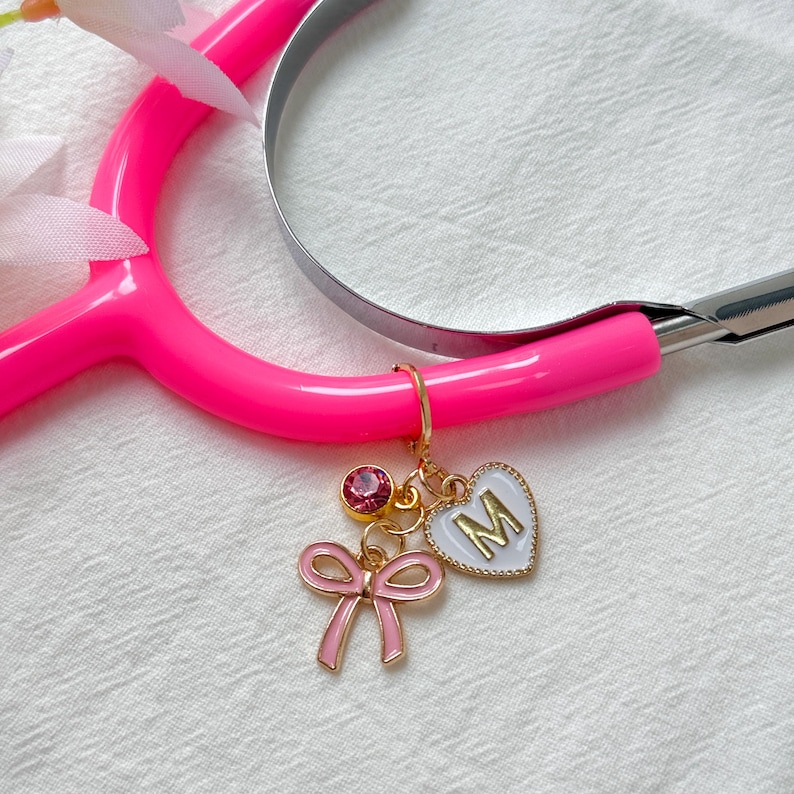 Puede incluir: Un estetoscopio rosa brillante con un dije dorado. El dije incluye un lazo rosa, una gema rosa y un coraz&oacute;n blanco con la letra "M". El estetoscopio est&aacute; sobre una superficie blanca texturizada.