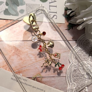 May include: A clear phone case with a gold heart-shaped clasp and a charm bracelet. The charm bracelet features various gold charms, including a red horse, a cherry, a bow, and a charm with the letter "K".