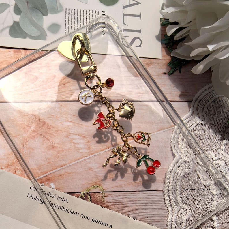 Puede incluir: Una funda transparente para tel&eacute;fono con un cierre dorado en forma de coraz&oacute;n y una pulsera de abalorios. La pulsera de abalorios presenta varios abalorios dorados, incluyendo un caballo rojo, una cereza, un lazo y un abalorio con la letra "K".