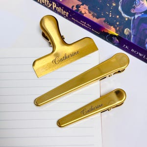 K&ouml;nnte beinhalten: Drei goldfarbene Metallclips in verschiedenen Gr&ouml;&szlig;en, angeordnet auf einer wei&szlig;en Oberfl&auml;che. Der gr&ouml;&szlig;te Clip ist ein Beutelclip, die anderen beiden sind kleinere B&uuml;roklammern. Der Name "Catherine" ist auf dem Beutelclip und einer der B&uuml;roklammern eingraviert.