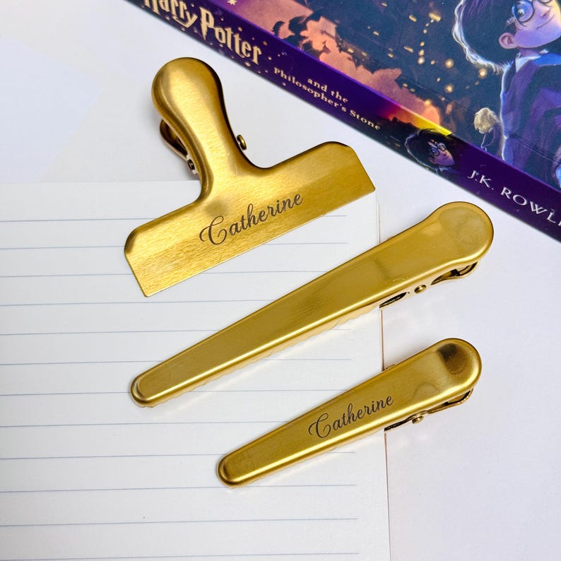 K&ouml;nnte beinhalten: Drei goldfarbene Metallclips in verschiedenen Gr&ouml;&szlig;en, angeordnet auf einer wei&szlig;en Oberfl&auml;che. Der gr&ouml;&szlig;te Clip ist ein Beutelclip, die anderen beiden sind kleinere B&uuml;roklammern. Der Name "Catherine" ist auf dem Beutelclip und einer der B&uuml;roklammern eingraviert.