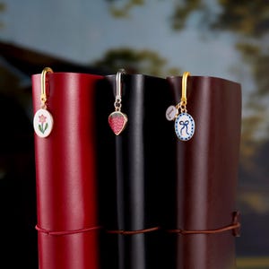 May include: Three decorative bookmarks with gold or silver hooks. One has a pink flower design, another a red strawberry, and the third a blue bow. The bookmarks are attached to leather-bound journals in red, black, and brown.