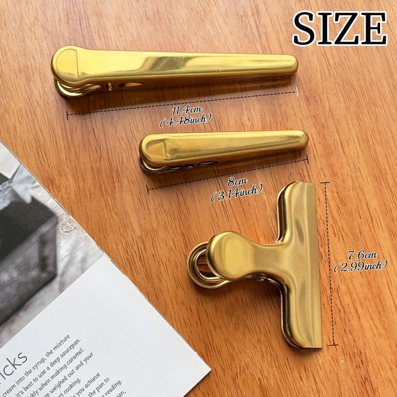 May include: Three gold-colored metal clips of varying sizes are displayed on a wooden surface. The largest clip measures 11.4 cm (4.48 inches) in length, the medium clip is 8 cm (3.14 inches), and the third clip is 7.6 cm (2.99 inches). The word "SIZE" is in the top right corner.