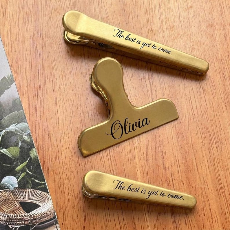 May include: Three gold-colored metal clips. Two clips have the text "The best is yet to come." The other clip has the name "Olivia" in cursive. The clips are on a wooden surface.