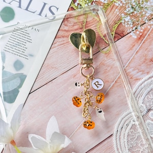 Könnte beinhalten: Ein goldfarbener Schlüsselanhänger mit herzförmigem Verschluss und einem Charm-Cluster. Der Cluster enthält orangefarbene Kürbis-Charms, einen weißen Geister-Charm und einen weißen runden Charm mit dem Buchstaben "R". Der Schlüsselanhänger ist an einer transparenten Handyhülle befestigt.