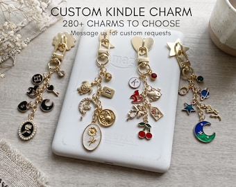 Könnte beinhalten: Vier goldfarbene Metallanhänger mit verschiedenen baumelnden Elementen wie Buchstaben, Blumen, Himmelskörpern und Tieren. Der Text lautet: CUSTOM KINDLE CHARM, 280+ CHARMS TO CHOOSE, Message us for custom requests. Die Anhänger liegen auf einem weißen E-Reader.