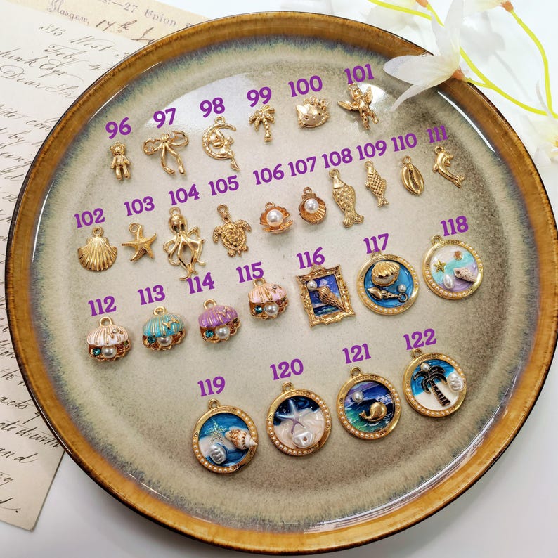 Puede incluir: Una colecci&oacute;n de abalorios y colgantes dorados, que incluyen conchas marinas, estrellas de mar y dise&ntilde;os celestiales. Algunos abalorios presentan perlas y detalles de esmalte de colores. Los abalorios est&aacute;n dispuestos en un plato decorativo.