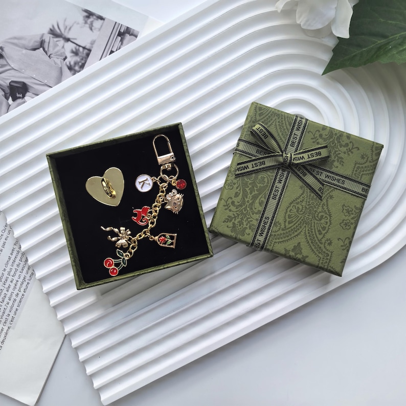 Puede incluir: Un pin dorado en forma de coraz&oacute;n y un llavero dorado con dijes, que incluyen cerezas, una rosa y un gato, se exhiben en una caja verde. Tambi&eacute;n hay una caja de regalo verde separada con una cinta "BEST WISHES".
