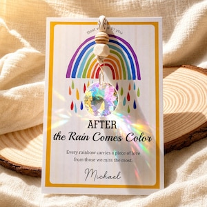 May include: A suncatcher ornament featuring a faceted crystal sphere hanging from a string of wooden beads, displayed on a white card with a rainbow graphic and the text: Best wishes to you, AFTER the Rain Comes Color, Every rainbow carries a piece of love from those we miss the most, Michael.