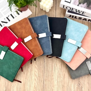 May include: A collection of colorful, leather-bound notebooks in various shades including green, red, brown, blue, black, light blue, pink, and gray. Each notebook features a silver clasp and a ribbon bookmark. The notebooks are arranged on a wooden surface.