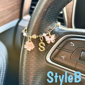 May include: A gold-colored car charm with a chain and multiple charms. The charms include a pink flower, a gold letter "S", and a pink butterfly. The charm is attached to a car steering wheel. The text "StyleB" is visible.