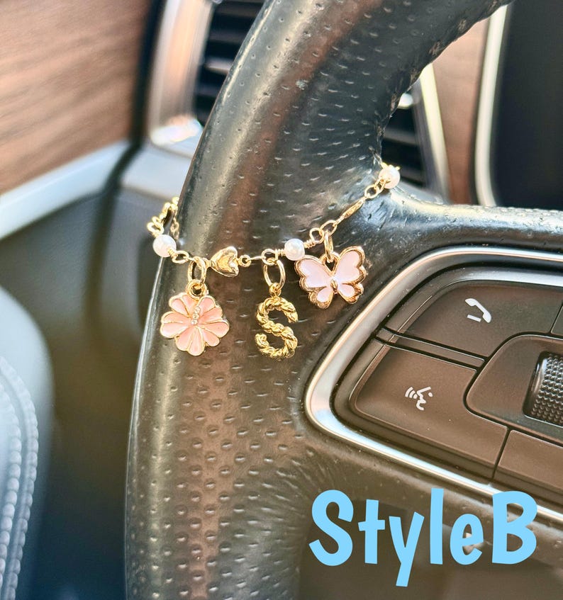 May include: A gold-colored car charm with a chain and multiple charms. The charms include a pink flower, a gold letter "S", and a pink butterfly. The charm is attached to a car steering wheel. The text "StyleB" is visible.