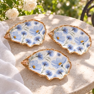 Puede incluir: Tres platos decorativos en forma de concha de ostra con un patrón floral azul pintado a mano y centros amarillos. Cada concha tiene un borde festoneado dorado y está personalizada con los nombres Lucy, Noah y Amelia en letras doradas.