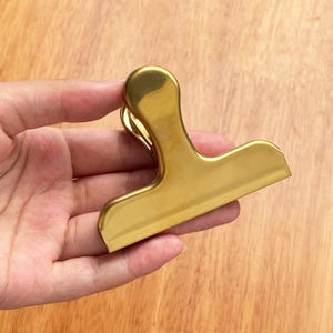 May include: A gold-colored metal bag clip with a rounded handle and a wide, flat clamping surface. The clip is held against a wooden surface, showcasing its shiny, metallic finish. This clip is designed for sealing bags.