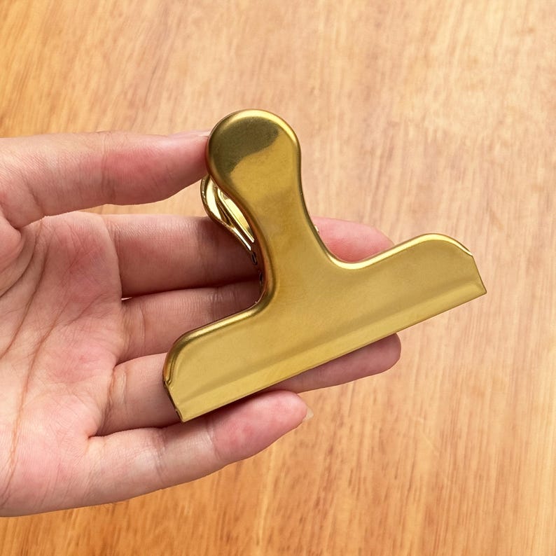 May include: A gold-colored metal bag clip with a rounded handle and a wide, flat clamping surface. The clip is held against a wooden surface, showcasing its shiny, metallic finish. This clip is designed for sealing bags.