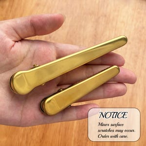 May include: Two gold-colored hair clips, one large and one small, are displayed. The clips have a smooth, shiny finish. The image includes a notice about minor surface scratches.