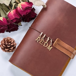 以下が含まれることがあります： 茶色の革製ジャーナル。ストラップ留めと「REDM」の文字を綴るゴールドカラーのチャームが付いています。背景にはドライレッドローズと松ぼっくりがあります。ジャーナルは閉じられています。
