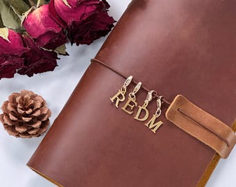以下が含まれることがあります： 茶色の革製ジャーナル。ストラップ留めと「REDM」の文字を綴るゴールドカラーのチャームが付いています。背景にはドライレッドローズと松ぼっくりがあります。ジャーナルは閉じられています。