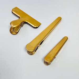 K&ouml;nnte beinhalten: Drei goldfarbene Metall-Beutelclips in verschiedenen Gr&ouml;&szlig;en. Der gr&ouml;&szlig;te Clip hat eine T-Form, w&auml;hrend die anderen beiden lang und rechteckig sind. Die Clips dienen zum Verschlie&szlig;en von Beuteln und zum Frischhalten des Inhalts.
