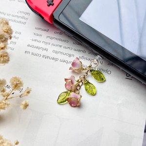 Pu&ograve; includere: Un ciondolo color oro con fiori rosa e bianchi e foglie verdi. Il ciondolo &egrave; attaccato a una spina di plastica trasparente, probabilmente per un dispositivo di gioco. Lo sfondo include un dispositivo di gioco e testo stampato.
