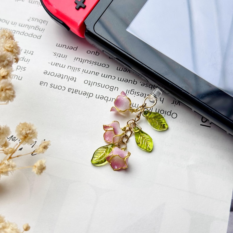 Pu&ograve; includere: Un ciondolo color oro con fiori rosa e bianchi e foglie verdi. Il ciondolo &egrave; attaccato a una spina di plastica trasparente, probabilmente per un dispositivo di gioco. Lo sfondo include un dispositivo di gioco e testo stampato.
