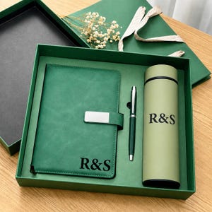 Könnte beinhalten: Ein grünes Geschenkset in einer passenden Box, bestehend aus einem Notizbuch mit silbernem Verschluss, einem Kugelschreiber und einer zylindrischen Flasche. Notizbuch und Flasche sind mit dem schwarzen Text R&S personalisiert. Ein kleiner Zweig weißer Trockenblumen liegt bei.