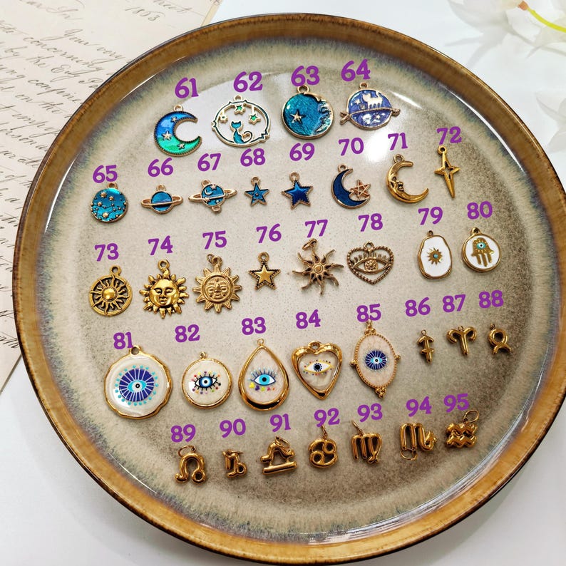 Puede incluir: Una colecci&oacute;n de colgantes dorados con varios dise&ntilde;os celestiales y simb&oacute;licos, incluyendo lunas, estrellas, soles y signos del zodiaco. Los colgantes se exhiben en un plato de cer&aacute;mica decorativo, mostrando una variedad de colores y texturas.