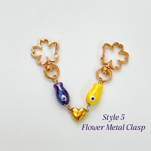 Op de afbeelding: Een goudkleurige metalen sluiting met een bloemdesign aan elk uiteinde, verbonden door een ketting. De ketting heeft een paarse en een gele tulpvormige bedel en een gouden hartbedel. De tekst "Style 5 Flower Metal Clasp" is zichtbaar.