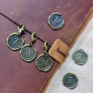 Puede incluir: Encantos iniciales de color bronce antiguo con cierres de langosta, con las letras F, W y N. También son visibles encantos iniciales adicionales con las letras V, E y J. Los encantos se muestran en un diario de cuero marrón.