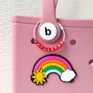 Könnte beinhalten: Eine rosa Tragetasche mit einem weißen kreisförmigen Anhänger mit dem Buchstaben "b" und dem Namen "CATHERINE" in roten Buchstabenperlen. Ein Regenbogen- und Sonnenanhänger ist an der Tasche befestigt.